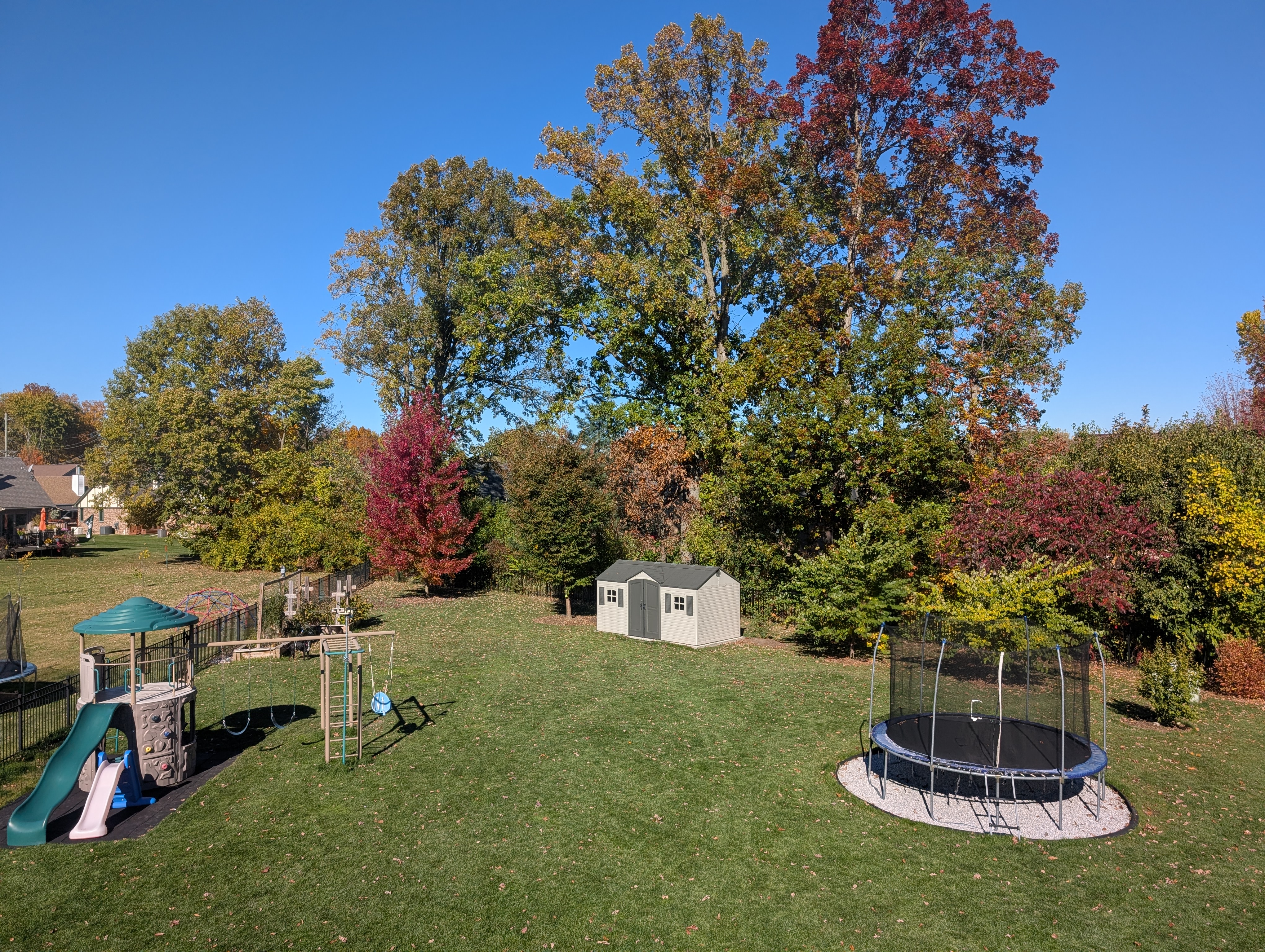 A sunlit backyard in autumn with a wooden playset, swing, and trampoline surrounded by trees beginning to turn orange and gold